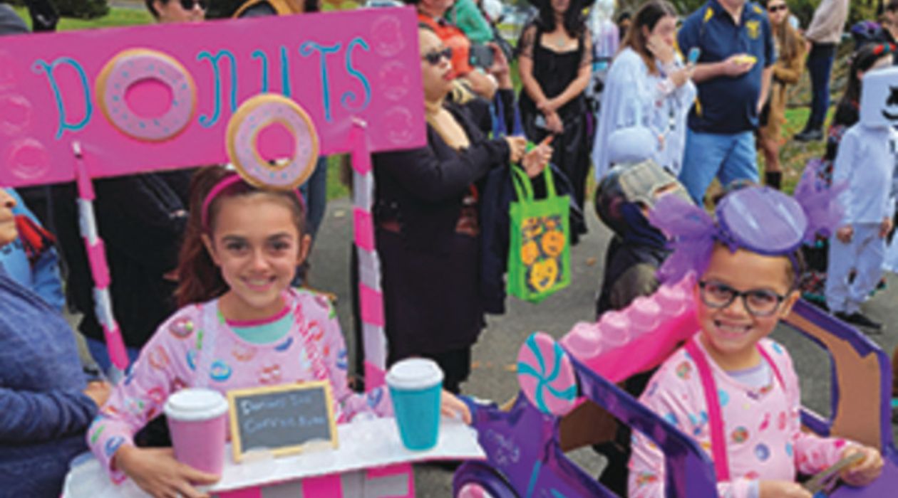 Two young kids in cute costumes smile as they enjoy the Wicked Wappingers Halloween Festival and Parade. One is dressed as a donut stand with a big pink sign adorned with donuts above her head and a countertop with brightly colored coffee cups in front of her. The other is dressed as a bright purple car decorated with pink and blue candies.